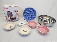 Photo showing entire lot including Dash treat maker box, large stainless steel bowl, blue slow feeder, and five small animal-themed ceramic bowls.