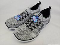 Pair of Skechers women's slip-on shoes showing grey and black knit uppers with tags attached, side view showing 'S' logo and 'Dual-Lite' label on sole