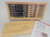 Six steak knives with black handles packed neatly in a pine wooden box with the lid showing the Wolfgang Puck logo, accompanied by a use and care booklet.