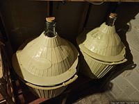 Two large 15 gallon glass carboys each with a beige plastic protective basket, placed on the floor.