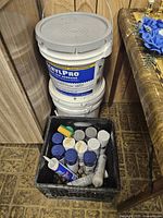 Black crate with multiple spray paint cans and caulk tubes inside, showing various colors and brands, next to two white plastic buckets of flooring adhesive stacked vertically.