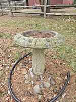 Photo showing full bird bath in yard with floral pattern rim and pedestal base surrounded by rocks and pine needles.