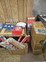 Image showing boxed artificial Christmas trees, several rolls of Christmas wrapping paper in a cardboard box, a red box, and other items on a table.