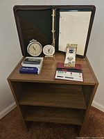 Wooden shelf with various items displayed on top including two vintage clocks, a checkbook cover with solar calculator, new wallet in box, and brown padfolio with notepad open inside.