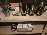 Shelf with vintage coffee pots including stovetop espresso maker, electric espresso maker with box, stainless steel percolator pots, and boxed ceramic accessories.