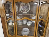 China cabinet with multiple white plates, bowls, tureen, and large decorative plate visible inside.