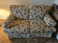 Front view of a two-seater loveseat with a floral fabric upholstery featuring cushions on armrests and back, beige carpeted floor beneath.
