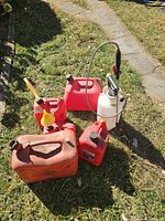 Five red plastic gas cans of various sizes arranged on grass along with one white garden sprayer with a pump handle and hose