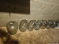 Seven glass carboys/demijohns lined up on a concrete floor against a cinder block wall, showing overall shape and color.