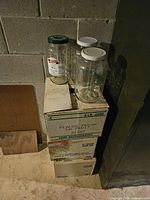 Four clear glass jars on top of cardboard boxes, two with white lids, one with a dark green lid, one without a lid, set against a concrete wall.