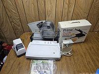 Photo shows the lot including an electric food slicer, electric vacuum sealer with vacuum bags, electric knife sharpener, and boxed Prima Pizelle Baker on a wooden table.