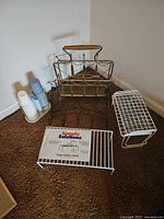 Group of racks including white metal wire shelf racks, a white plastic dispenser holder for bottles, and the brass tone metal magazine rack with wooden handles visible behind.