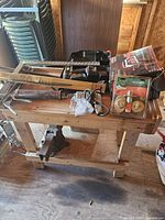 Wide view of wooden workbench with attached metal vice, cordless drill in case, battery charger box, and unopened deadbolt lock set on workbench.
