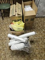 Boxes and plastic bags containing new disposable cups and plates in white color, some visible packaging with quantities.