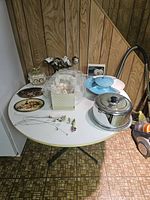 Photo of a white round table with various items including California pottery lazy susan serving set in blue and white, metal domed cake plate, ceramic item in bubble wrap, doll box, and framed pictures.