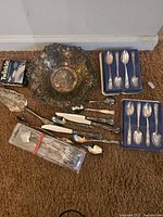 Full view of metal decorative bowl, two boxed sets of commemorative spoons, sealed set of flatware, assorted pie servers, knives and spoons laid on carpet