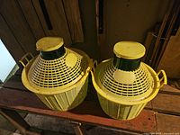 Two glass carboys on a wooden shelf, each with a yellow plastic basket-style protective cover and yellow lids.