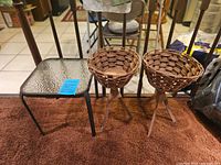 Photo showing the small outdoor table with metal frame and textured glass top alongside two rattan woven plant stands with tripod legs, placed on a brown surface indoors.