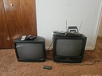 Photo showing two small CRT televisions side by side on carpeted floor, one with remote on top and one TV/VCR combination with remotes and rabbit ear antennas.