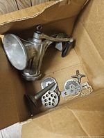 Top perspective showing vintage metal clamp-on device with crank handle and multiple metal discs and attachments inside a cardboard box.