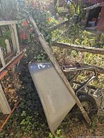 Wheelbarrow leaning against a wooden fence outdoors, showing full length including handles and tray in natural light.
