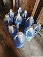 10 plastic jugs of blue windshield washing fluid arranged on a wooden shed floor. Bottles are translucent and some dusty, with clear screw caps and handles visible.