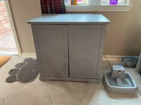 Gray wooden cat house/litter box enclosure shown from front with two closed doors and a pet water fountain beside it.