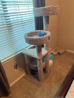 Full view of beige carpeted multi-level cat tree with visible wear and damage. Shows top round bed, platforms, and scratching posts.