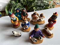 Top angled view of all clay nativity scene figurines arranged on white surface with leafy plants in background