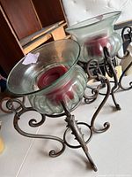 Two matching hurricane candle holders with clear glass, red candles, and ornate iron stands on a white tufted background.