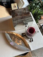 Overall view of the metal hurricane candle holder with red candle, metal oblong tray, and glass display stand on a surface with additional plants and boxes in background.