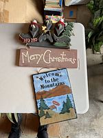 Photo showing two wooden hanging signs: a 'Merry Christmas' holiday sign with birds and tree, and a mountain-themed 'Welcome to the Mountains' sign