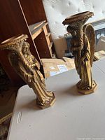 Two golden angel candle stick holders standing upright on a table, showing front and sides of both items.