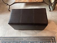 Top view of one leather storage bench, rectangular, dark brown with stitched pattern on top, placed on tiled floor.