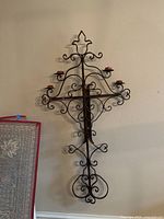 Photo of one wrought iron wall hanging cross with scrolled arms and candle holders against a light wall near a chair and blanket.