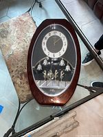 Full view of Menet oval wall hanging clock on glass table, showing wooden frame and clock face