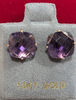 Close-up photo of the amethyst earrings resting on a grey velvet holder stamped with '14KT GOLD' in gold lettering.