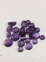 Top-down view of approximately 20 small polished genuine amethyst gemstones in various shapes and sizes with purple hues.