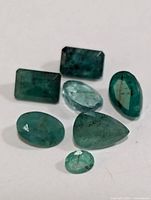 Photo showing six faceted genuine emerald gemstones in various cuts and shades of green with natural inclusions.