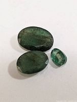Three genuine emerald gemstones displayed on white background, showing color and cut.