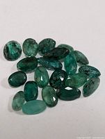 Top-down view of approximately 22 loose genuine emerald gemstones showing various cuts, green hues, and natural inclusions.