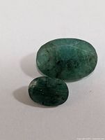 Two oval genuine emerald gemstones placed on a white surface, showing size and color variations with natural inclusions visible.
