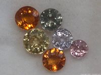 Image of six loose round cut fancy color sapphire gemstones in orange, yellow, gray, purple, and pink colors laid on white background.