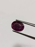 Close-up image of an oval-shaped ruby gemstone held by tweezers, showing the stone's facets and internal inclusions against a plain background.