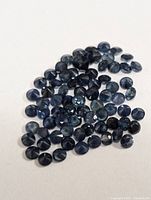 Loose pile of small, round faceted genuine sapphire gemstones on a white surface shown from above.
