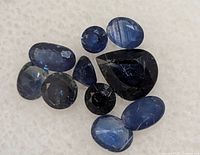Top down photo of 10 loose sapphire gemstones in various shapes and cuts on a light background showing their blue color and natural inclusions.