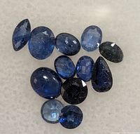 Top view of 12 variously cut and sized natural sapphire gemstones in shades of blue and dark blue, arranged loosely on a textured surface.