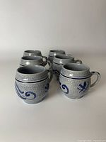 Front angled view of all six blue pottery mugs arranged in a group showing hand-painted decorative designs and bulbous shape.