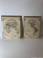 Two antique maps of Eastern and Western Hemispheres shown side-by-side standing up against a wall, each in a plastic sleeve.