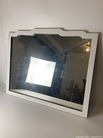 Front perspective view of the antique white painted wood framed mirror, showing its rectangular shape with decorative top edge and worn white paint.